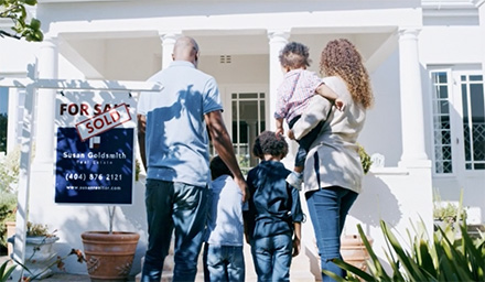 Decorative Image: A young family standing in front of the new home they purchased with the for sale/ sold sign next to them.