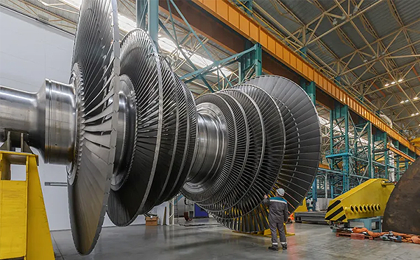 a worker standing in front of a large metal power turbine in a factory. Or a worker stands in front of a massive metal power turbine in factory OR the back of a worker standing in front of large metal power turbine.
 